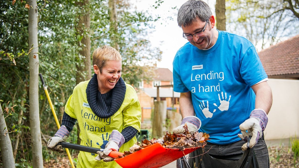 A smiling man and woman from Skanska putting leaves into a bag as part of a volunteering day